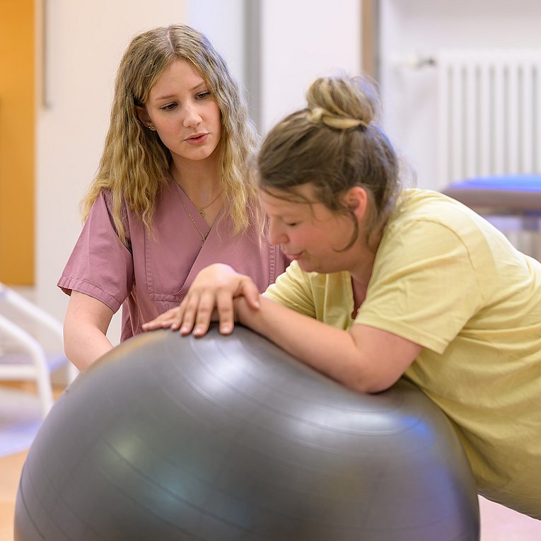 Eine Hebamme begleitet eine in den Wehen liegende Frau, welche sich auf einen Gymnastikball stützt. Beide atmen ruhig ein und aus.