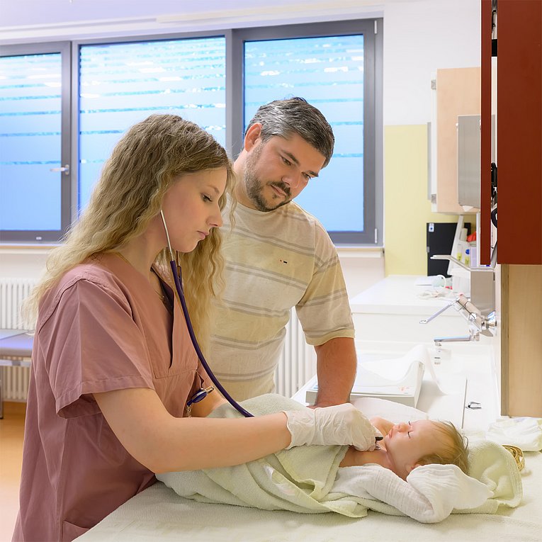 Eine Hebamme untersucht ein Baby, welches auf einem Wickeltisch liegt. Der Vater des Kindes schaut ihr dabei aufmerksam zu.